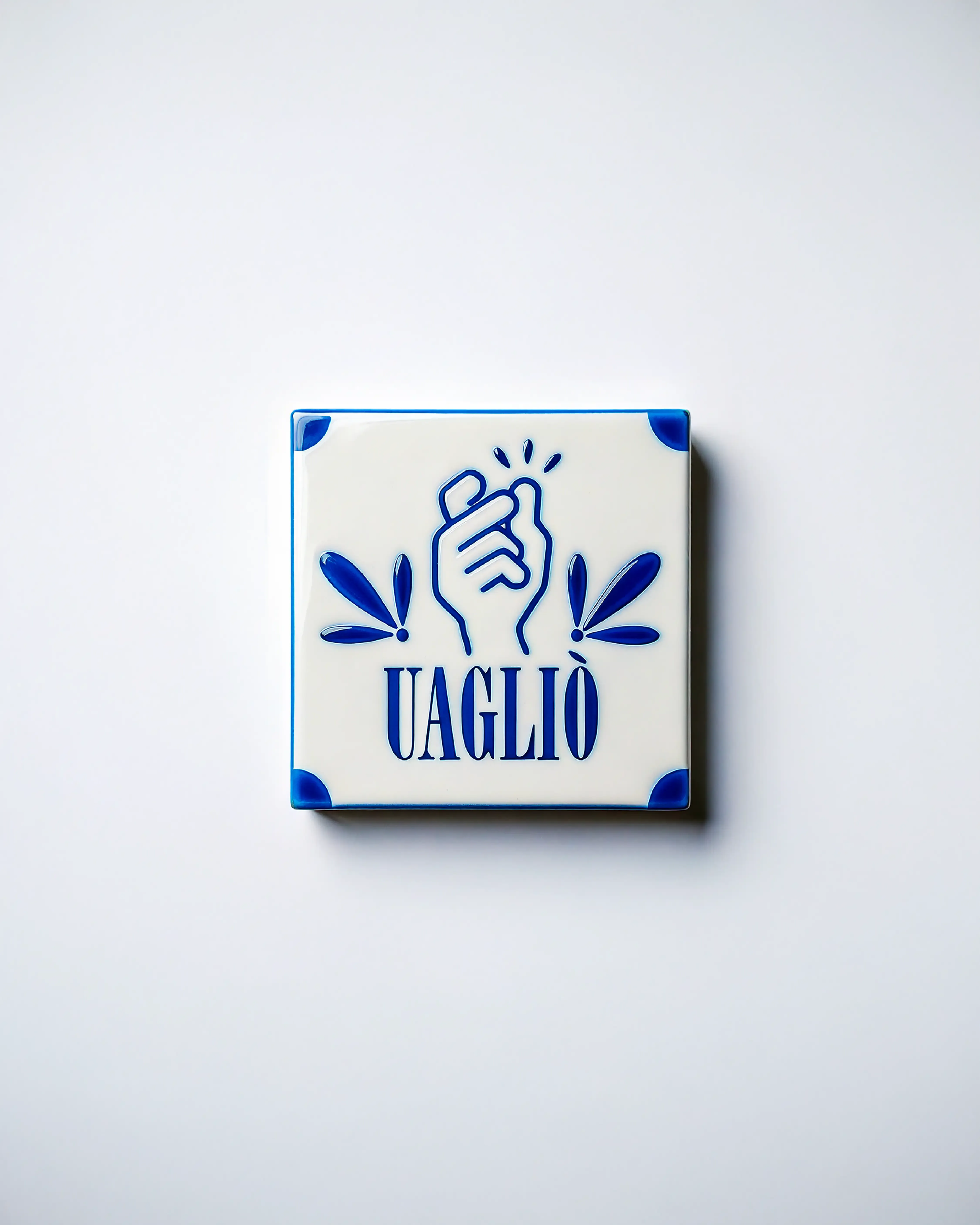 Piastrella in maiolica bianca e blu con la scritta Uagliò e una mano che schiocca le dita, motivo grafico di Pistill'